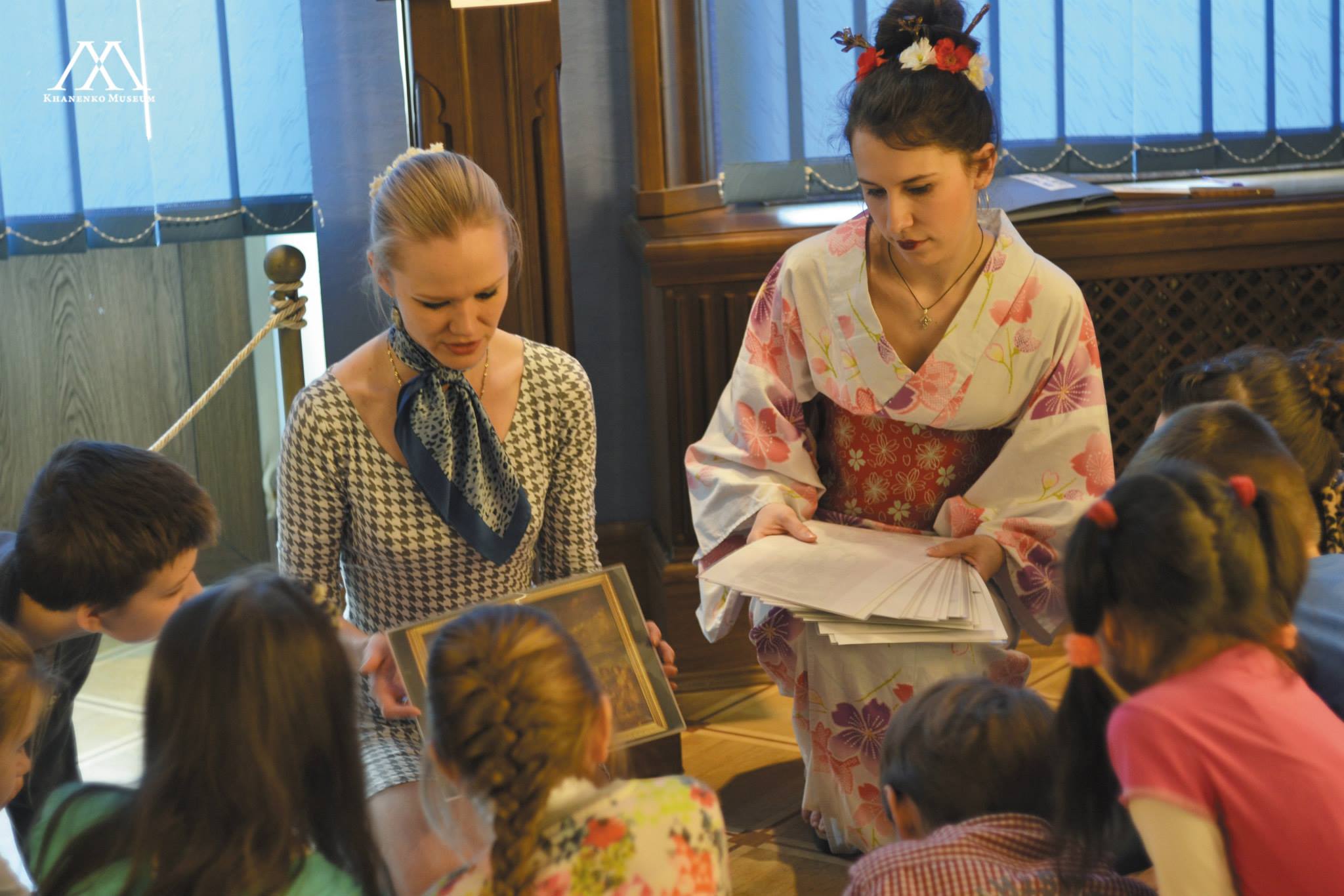 Photo. Two presenters, one  in a Japanese kimono, showing cards with illustrations to a group of children