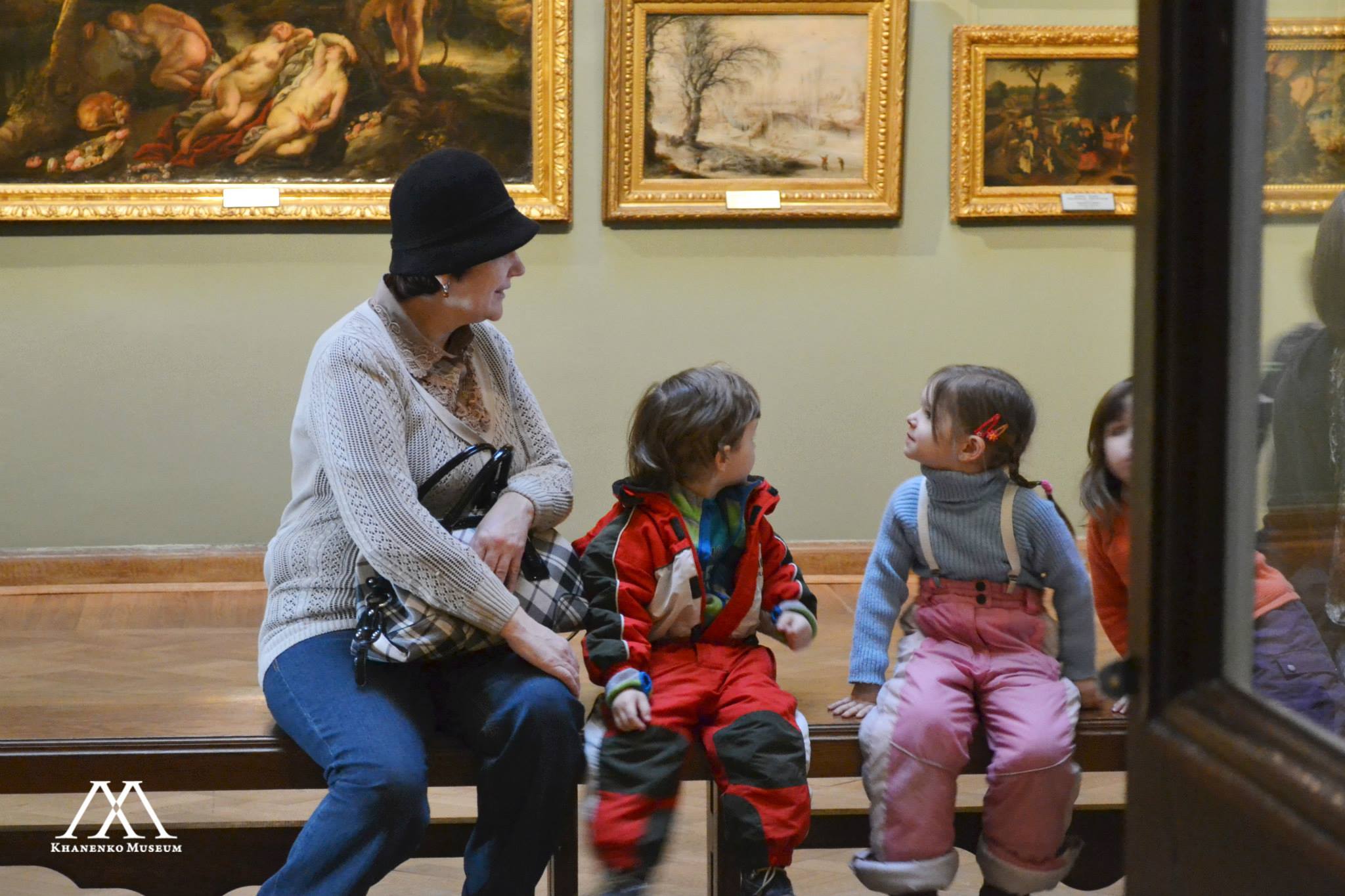Photo. A woman and three children are looking at the paintings around the gallery hall.